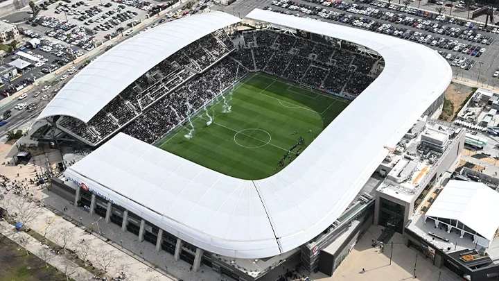 An aerial photograph showing the Stadium in Exposition Park from above, framed by two parking lots around the perimeter. Three plumes of white smoke rise from the football pitch with a crowd of spectators in the background.
