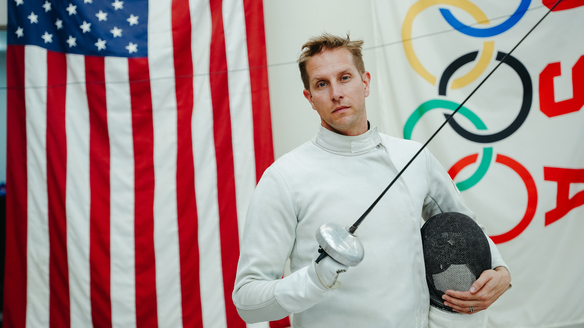 Cody Mattern standing wearing fencing gear in front of USA and IOC flags holding a fencing helmet and fencing weapon