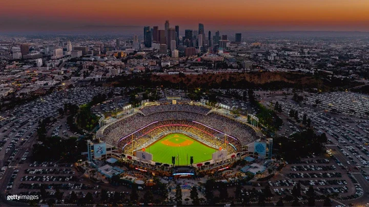 An aerial photograph of Dodger Stadium at sunset. The field of Dodger Stadium is illuminated while fans are seated. The parking lot surrounding the stadium is filled with cars and the downtown Los Angeles skyline is in the background. 